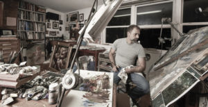 Donato Giancola, a man with short-cut dark hair and goatee, wearing a grey t-shirt and jeans, sitting in the midst of an artist's studio, with finished and in-progress paintings and paint supplies around him.