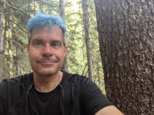 Kevin, a white man with short bright blue hair, wearing a black t-shirt, outside in a Pacific Northwest forest and surrounded by tall trees.
