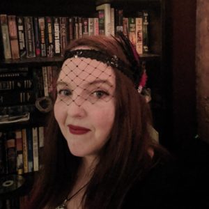 Michelle Morrell, a white woman with long straight dark brown hair, wearing a headband with veil and feathers and a black top, standing in front of a full bookcase.