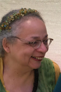 Nisi Shawl, a Black woman with grey hair, wearing glasses, a flowered hairband and a green top, smiling.