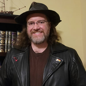 Richard Fife, a white man with medium-length brown hair and greying beard, wearing a leather hat, glasses, leather jacket with a progress pride flag pin, and brown shirt, standing in front of a bookcase.