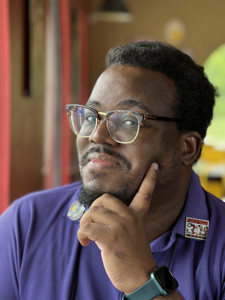 Brandon O'Brien, a Black man with short hair and goatee, wearing glasses and a purple polo shirt, with his chin on his hand and looking at the camera with a sly smile.