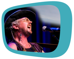 Alexander James Adams, a white man with long brown and grey hair in a ponytail, wearing a black sleeveless shirt and cowboy hat, plays a guitar and sings into a microphone.