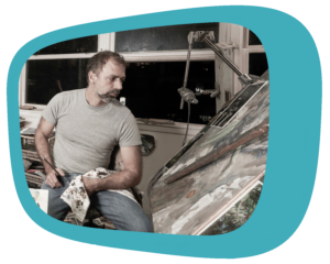 Donato Giancola, a man with short-cut dark hair and goatee, wearing a grey t-shirt and jeans, sitting in the midst of an artist's studio, with finished and in-progress paintings and paint supplies around him.