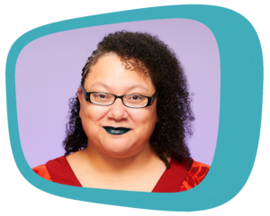 K Tempest Bradford, a Black woman with curly black hair, wearing glasses, black lipstick, and a red and orange top, smiles at the camera.