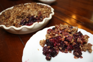 A white bowl filled with blackberry crisp sits next to a white plate with a serving of crisp, with deep red and purple berries covered by browned oats.