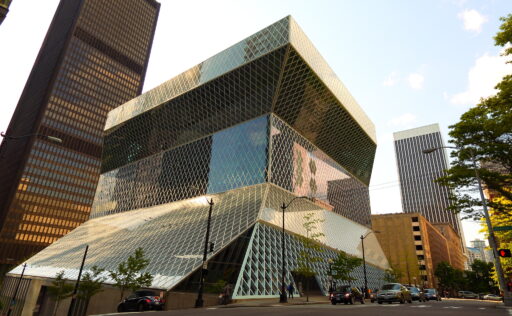 A large building, vaguely anvil-shaped from the angle shown, glass-clad behind a diagonal grid pattern, in the midst of more traditional blocky buildings in downtown Seattle, shown from street level on a sunny afternoon.