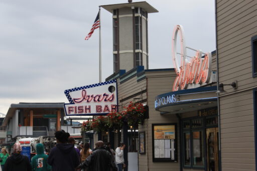 The outside of Ivar's Fish Bar on the Seattle Waterfront, with people in hoodies and jackets outside on a grey Seattle day. Baskets of red flowers hang underneath the neon Ivar's Fish Bar sign. A second neon sign over another door says Ivar's Acres of Clams.