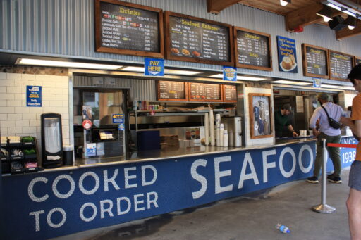 Inside Ivar's, menu boards line the wall above an ordering counter with people standing in line. Underneath the counter blue and white tiles spell out "Cooked to order seafood since 1938".