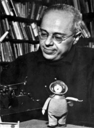 Black and white photo of Stanisław Lem, a bald white man wearing wire-rim glasses and a dark shirt, sitting at a table in front of a full bookcase, with a typewriter and small cosmonaut doll in front of him.