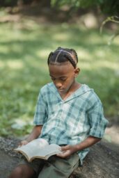 A Black boy with braided hair sits on a log in a field on a sunny day reading a book.