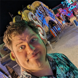 Keith Jewell, a whit man with short curly brown hair and muttonchops, wearing a green and black patterned shirt, standing in front of what appears to be a carnival ride sign.