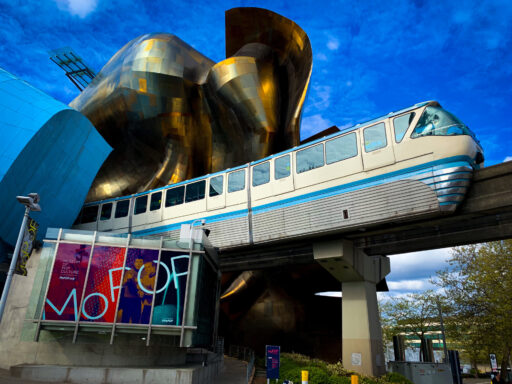 The Seattle monorail's blue train, white and silver with blue highlights, emerges on its elevated track from the blue and gold organic curves of the Museum of Pop Culture under a blue summer sky with wispy white clouds.