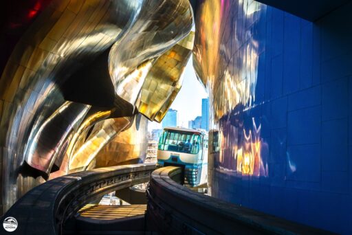 The Seattle monorail's blue train passing through the Museum of Pop Culture, a bright blue flat wall to the right reflecting the wavy, organically curved shiny gold overhanging wall to the left, with Seattle skyscrapers visible in the distance.