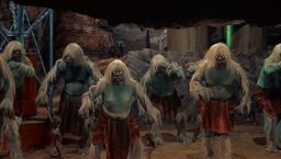 A group of Morlocks, light blue-skinned creatures with bestial faces and long white hair on their heads, arms, and legs, wearing reddish loincloths, in a cave with some metal scaffolding visible to one side.