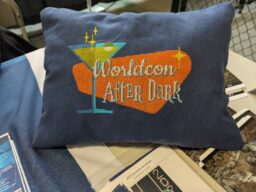 Cross stitch words "Worldcon After Dark" with a martini glass and stars, with a 1960s aesthetic in shades of orange, made into a pillow.