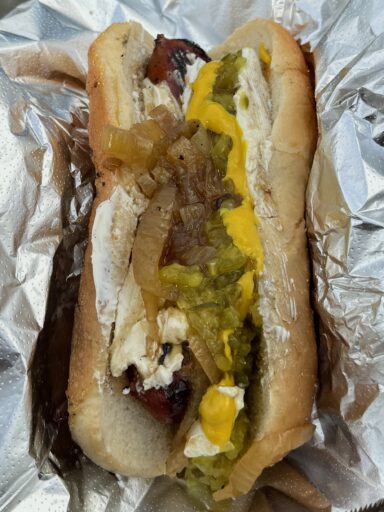 A hot dog covered in cream cheese, sautéed onions, relish, and mustard, in an open foil wrapper.