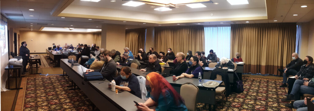 Panoramic photo of a hotel conference room with a large group of people sitting in rows at tables as a single person stands at the front of the room in front of a projection screen.