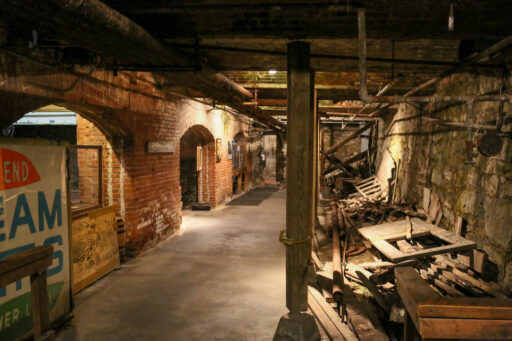 A concrete walkway through a basement with a brick wall with arched openings on the left side, and a jumble of discarded wooden beams and doors piled on the right side by an old stone retaining wall.