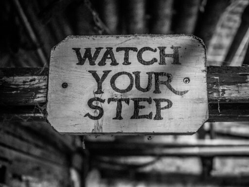 Black and white photo of a wooden handpainted sign reading Watch Your Step on an old, cobwebby support beam.