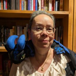 Mary Soon Lee, a woman with greying hair, wearing glasses and a white top, standing in front of a bookcase with a blue and black plush dragon draped across her shoulders.