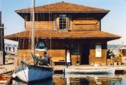 A two-story wooden building on a pier with a rowboat and a sailboat moored in front of it.