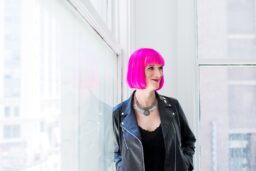 Charlie Jane Anders, a white woman with bright pink hair wearing a black top and black leather jacket, leans against a window in a white room.