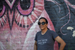 Bryan Thao Worra, a Lao man with brown skin and long straight black hair pulled back in a ponytail, wearing opaque goggles and a grey t-shirt that says 'I am my ancestors' wildest dreams', stands in front of a brick wall painted with a mural of a pink owl.