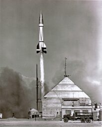 A V2 rocket launches from behind a metal shed with an army truck parked in front in 1949.