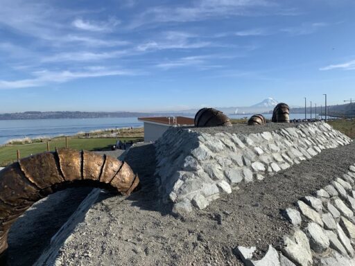 Sculptures of sandworms going in and out of stone and dirt platforms by the water of the Puget Sound with Mt. Rainier (Tahoma) visible in the distance under blue skies on a clear day.