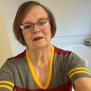 Sheril, a woman with straight brown hair, with red horn rim half frame glasses, wearing a grey, maroon, and yellow t-shirt.