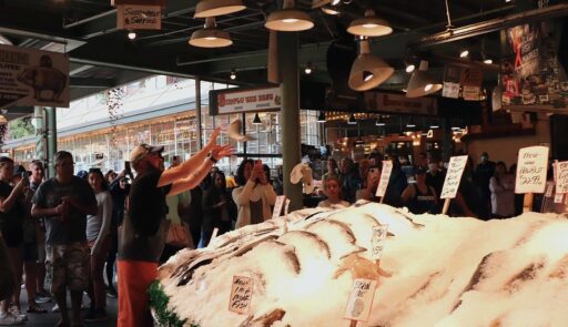 A fishmonger throws a large fish over a display of other fish laid out on ice as tourist look on and take photos.