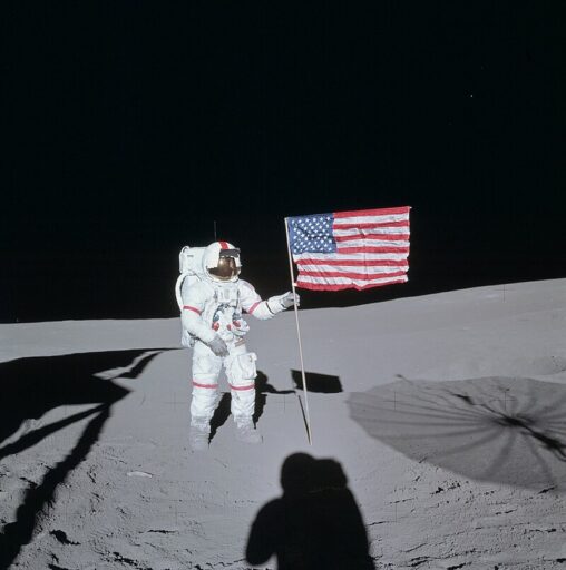Shepard in a white spacesuit with red stripes, standing on the surface of the moon and holding the American flag.