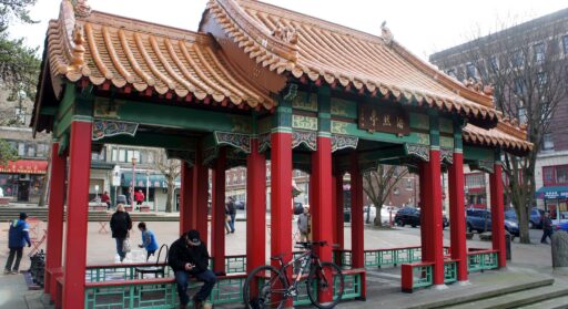A Chinese-style shelter with red poles, jade green accents, and sloped tan tiled roof, with adults and children visible in the plaza of Hing Hay Park behind the shelter.