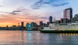 A white and green ferry docked at the Seattle port at sunset, with the Seattle skyline behind it, including the waterfront ferris wheel and the Space Needle in the distance.
