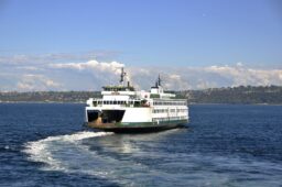 A green and white ferry trailing its wake as it heads out into the sound, with tree-covered land visible across the water.