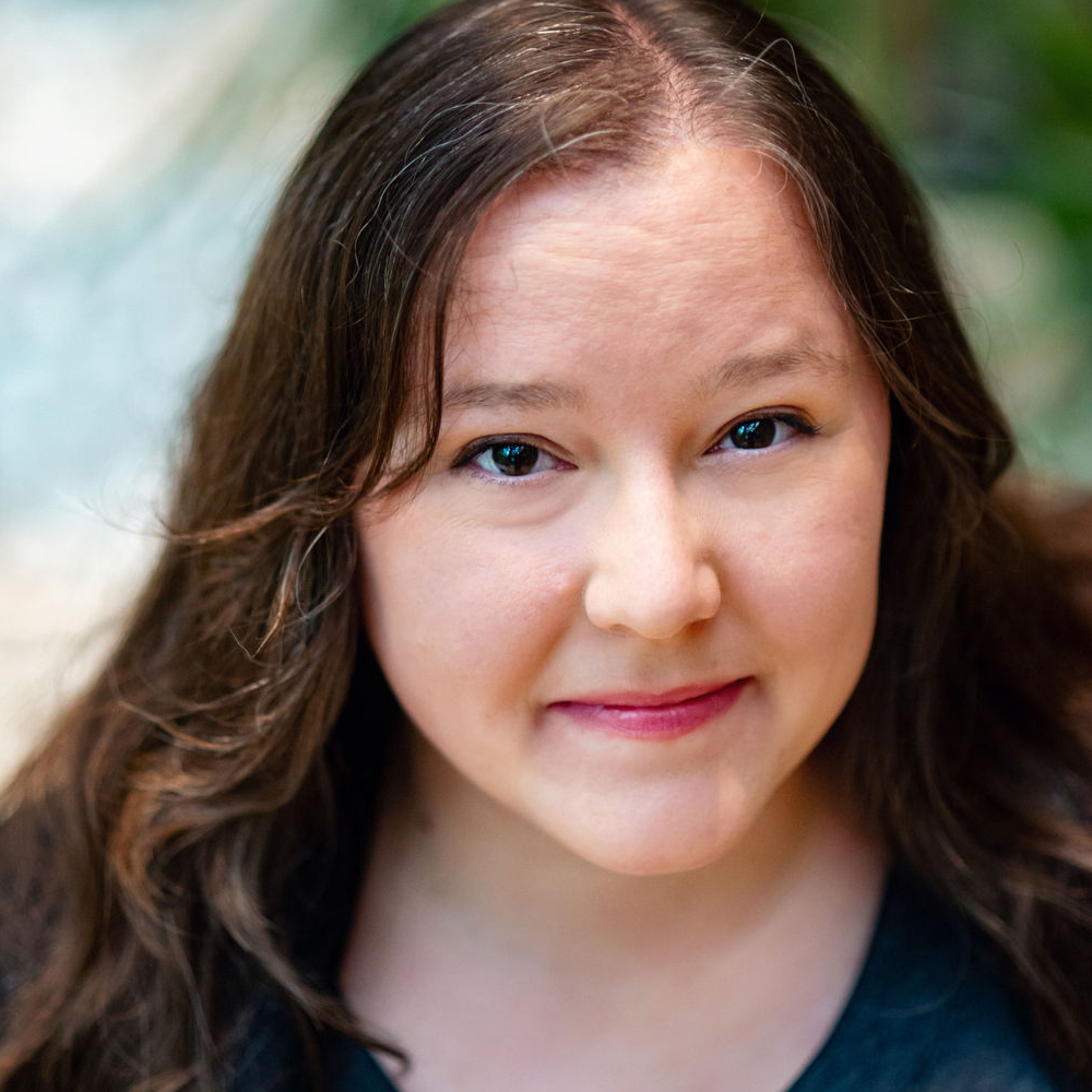A headshot of multidisciplinary storyteller A L Raden smiling up at the camera. She has long brown hair, brown eyes, and is wearing a gray shirt.