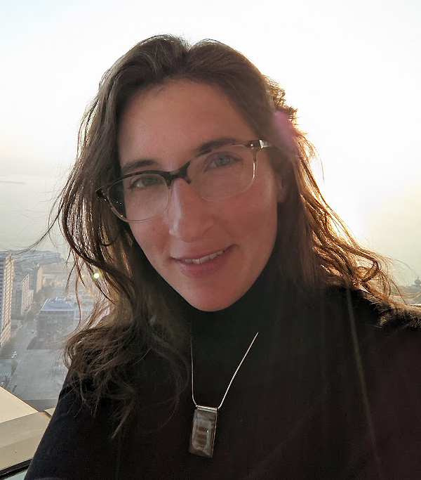 A woman with white skin and large rimmed glasses smiles at the camera with bright sunlight behind her. She is wearing a black turtleneck shirt and wears a silver necklace with a rectangular picture jasper pendant.