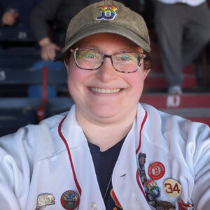 Gail Terman Gail Terman, a white woman with glasses, wearing a white and red baseball jersey with buttons and a baseball cap with a rainbow B logo, smiling and sitting in bleachers.