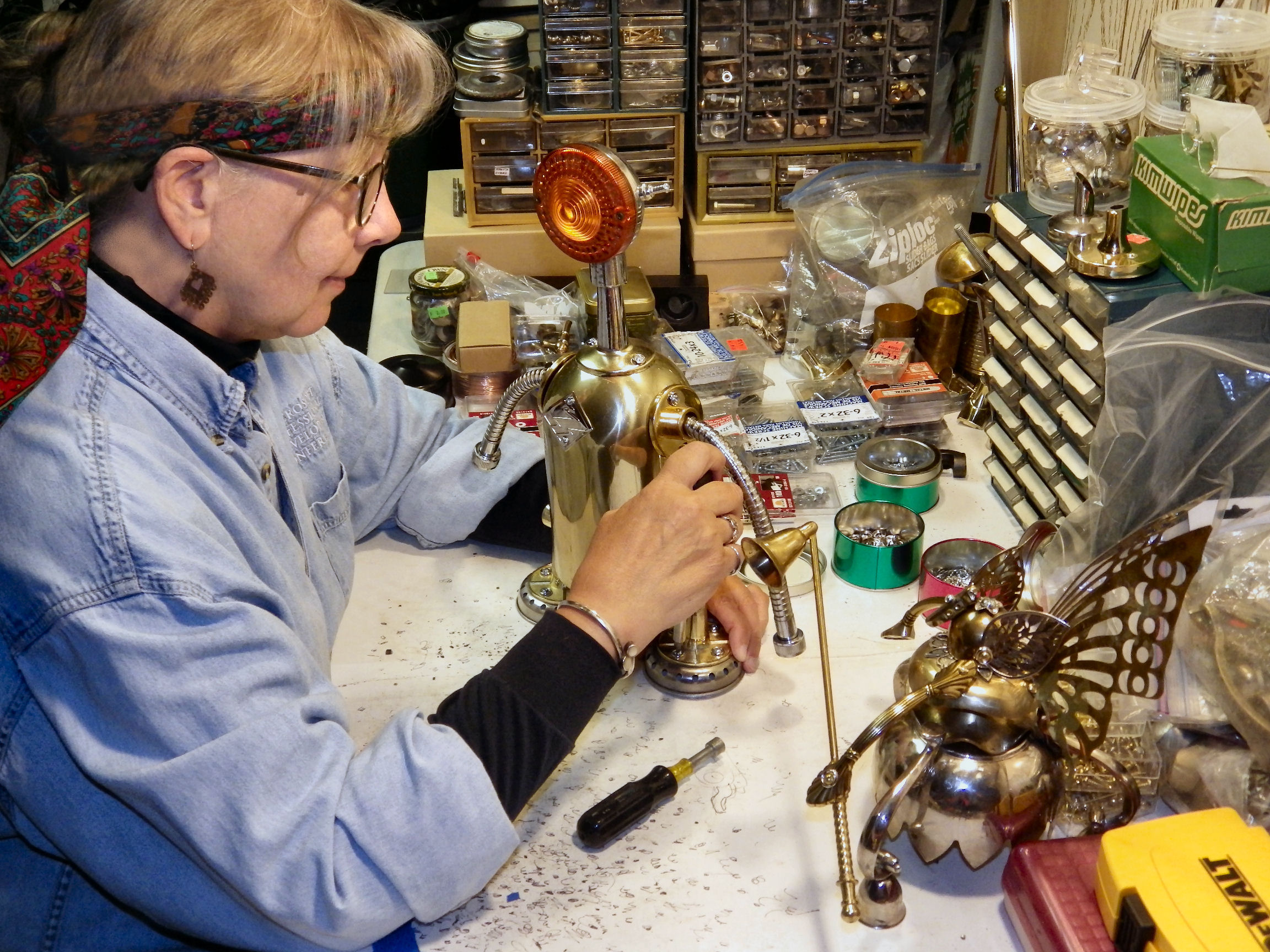Profile shot of Lynne (wearing her usual bandana around her head) working on constructing a Bot. The table is a mess of miscellaneous metal pieces. Across the back and side of the table are stacks of drawer cases containing screws and other metal bits used in construction. In the foreground are two finished Bots, a Retro robot and a fairy.