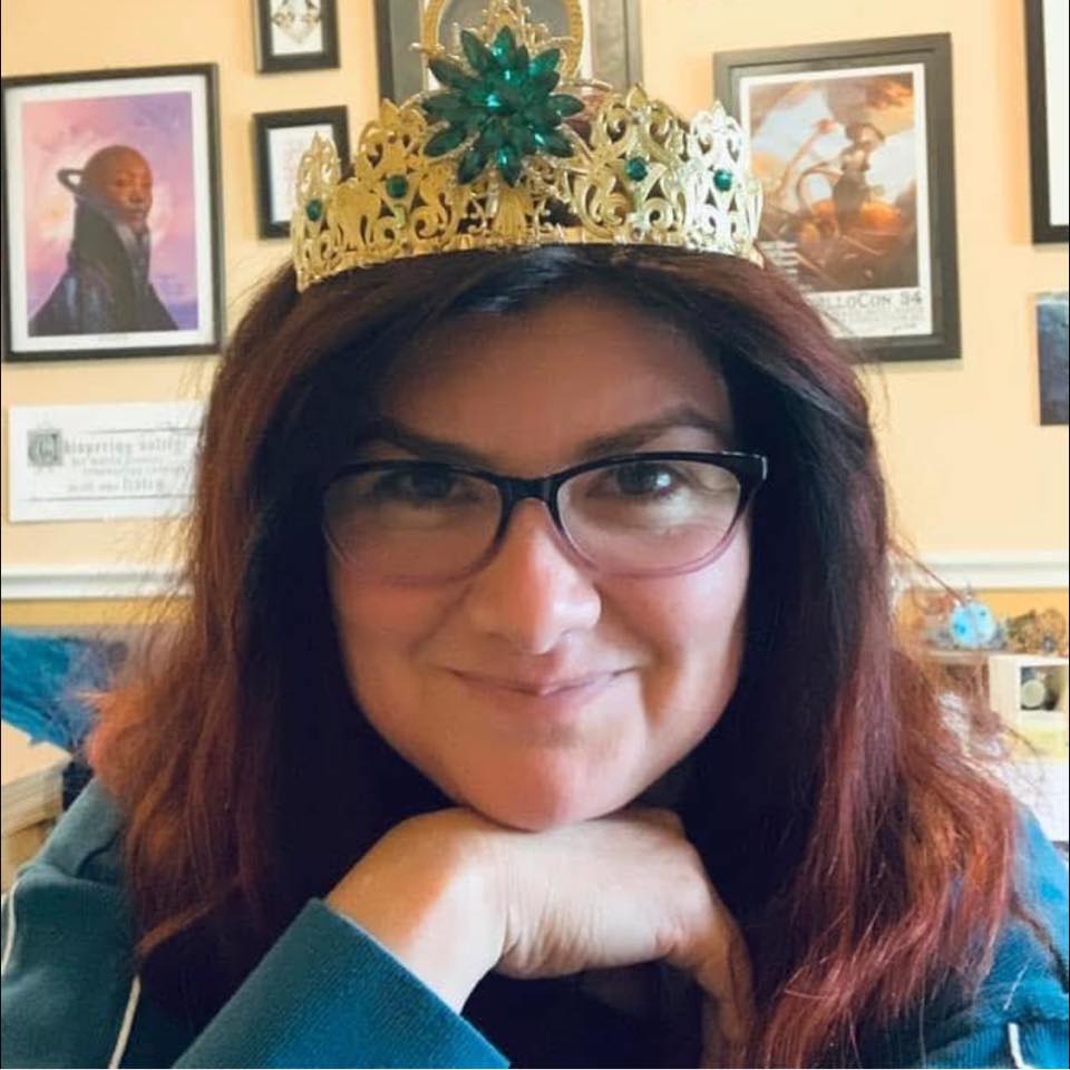 A picture of a brown haired woman leaning on her hand wearing a gold and green tiara.