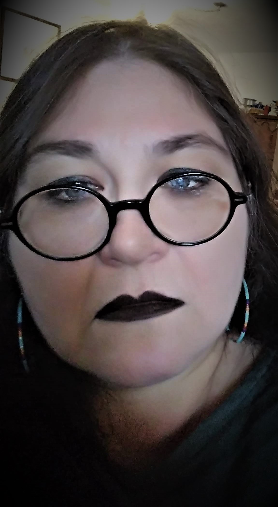 A light skinned woman with long dark hair, black lipstick, and round black glasses.