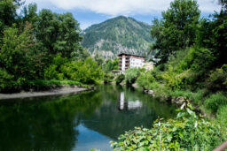 A quiet river flowing between forested banks, with a Bavarian-style hotel visible in front of a mountain hillside.