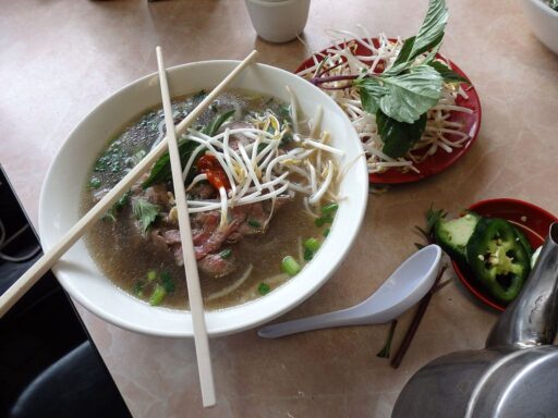 A white bowl of phở with broth, meat, noodles, and bean sprouts, with chopsticks crossed over the top, next to a red plate with extra bean sprouts.
