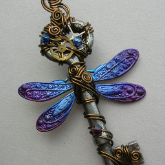 A cropped image of a wire wrapped antique key with brass and silver gears in the head, dragonfly wings that shade from light blue near the body to purple at the tips, matching crystal beads, and loops and spirals of antique brass colored wire.