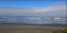 Waves of the Puget Sound breaking against a a sandy beach under cloudy blue skies.