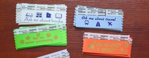 Photo of ribbons that adhere to badges with a peel-off adhesive strip. The badges shown say 'Ask me about books' in a blue font on a white background with several images of books. The second reads 'Ask me about travel' in dark blue on a light blue background with icons for a bus, a road, and an airplane beneath. The third shows a green ribbon with gold font that says 'Ask me about gardening' with five floral images floating above. The last shows an orange ribbons with a gold font that reads 'Ask me about sports' and has a football helmet, sports whistle, and basketball hoop icons.